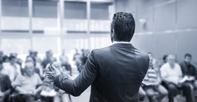 Rear view of speaker giving a talk at business meeting. Audience in the conference hall. Business and Entrepreneurship concept. Black and white blue toned image.
