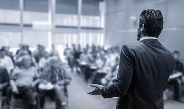 Rear view of speaker giving a talk at business meeting. Audience in the conference hall. Business and Entrepreneurship concept. Black and white blue toned image.