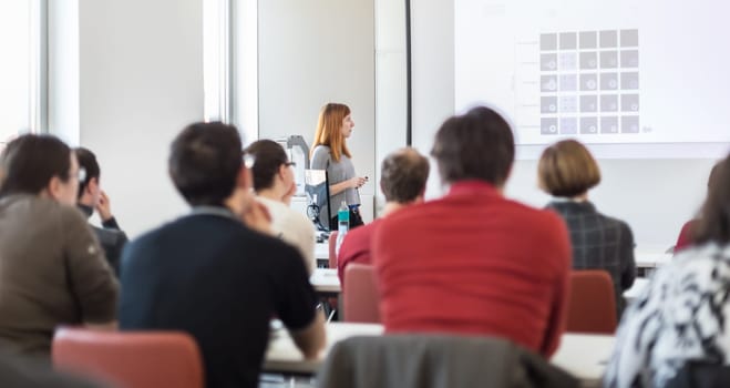 Female speaker giving presentation in lecture hall at university workshop. Audience in conference room. Rear view of unrecognized participant in audience. Scientific conference event.