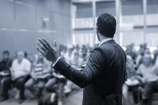 Rear view of speaker giving a talk at business meeting. Audience in the conference hall. Business and Entrepreneurship concept. Black and white blue toned image.