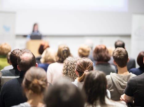 Female speaker giving presentation in lecture hall at university workshop. Audience in conference hall. Rear view of unrecognized participant in audience. Scientific conference event.