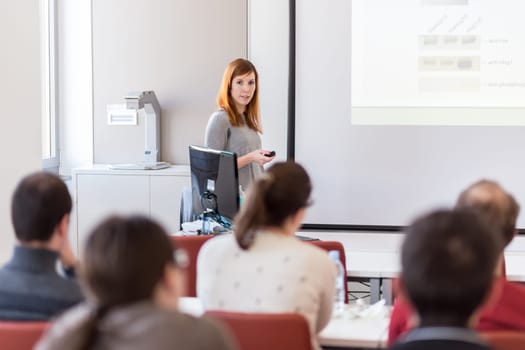 Female speaker giving presentation in lecture hall at university workshop. Audience in conference hall. Rear view of unrecognized participant in audience. Scientific conference event.