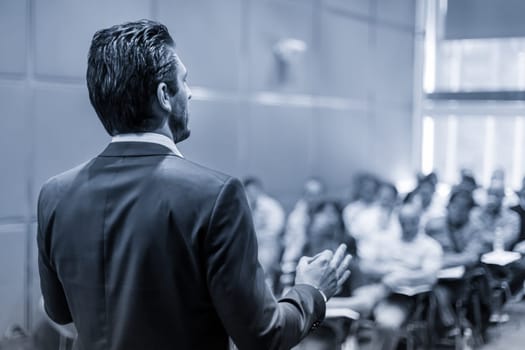 Rear view of speaker giving a talk at business meeting. Audience in the conference hall. Business and Entrepreneurship concept. Black and white blue toned image.