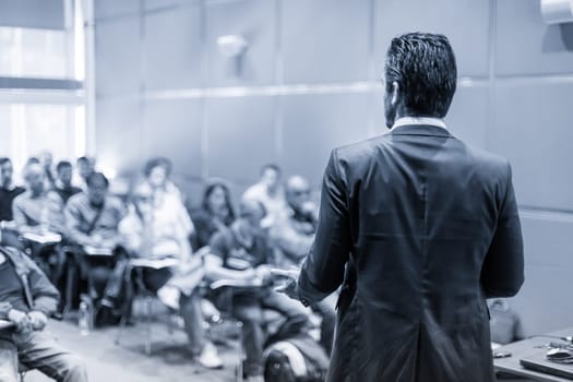 Rear view of speaker giving a talk at business meeting. Audience in the conference hall. Business and Entrepreneurship concept. Black and white blue toned image.