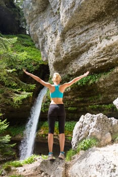 Active woman raising arms inhaling fresh air, feeling relaxed and free in beautiful natural environment under Pericnik waterfall in Vrata Valley in Triglav National Park in Julian Alps, Slovenia.
