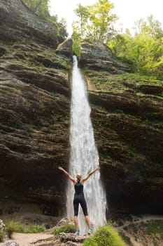 Active woman raising arms inhaling fresh air, feeling relaxed and free in beautiful natural environment under Pericnik waterfall in Vrata Valley in Triglav National Park in Julian Alps, Slovenia.