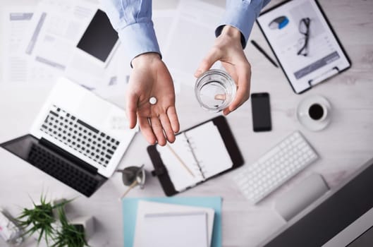 Office worker taking a pill and holding a glass of water hands close up with desktop on background, top view