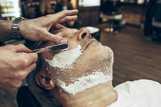 Close-up side top view handsome senior bearded caucasian man getting beard grooming in modern barbershop. Hairdresser serving client, making beard haircut using straight razor. Barber shop concept.