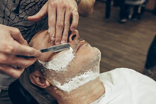 Close-up side top view handsome senior bearded caucasian man getting beard grooming in modern barbershop. Hairdresser serving client, making beard haircut using straight razor. Barber shop concept.