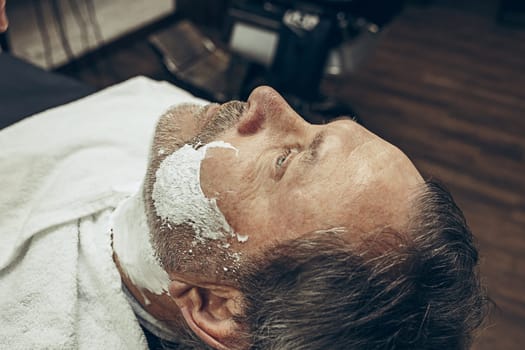 Close-up side top view handsome senior bearded caucasian man getting beard grooming in modern barbershop. Hairdresser serving client, making beard haircut using straight razor. Barber shop concept.
