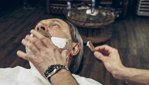 Close-up side top view handsome senior bearded caucasian man getting beard grooming in modern barbershop. Hairdresser serving client, making beard haircut using straight razor. Barber shop concept.