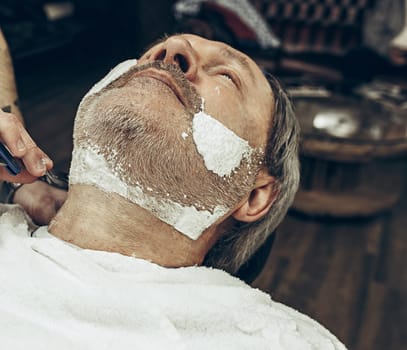 Close-up side top view handsome senior bearded caucasian man getting beard grooming in modern barbershop. Hairdresser serving client, making beard haircut using straight razor. Barber shop concept.