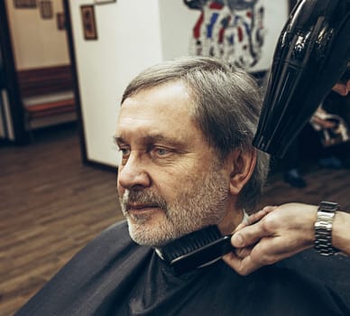 Close-up side view portrait of handsome senior bearded caucasian man getting beard grooming in modern barbershop. Hairdresser serving client, making beard haircut using shaving machine. Barber shop concept.