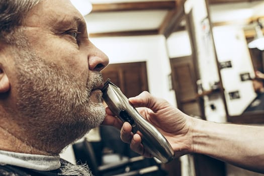 Close-up side profile portrait of handsome senior bearded caucasian man getting beard grooming in modern barbershop. Hairdresser serving client, making beard haircut using shaving machine. Barber shop concept.