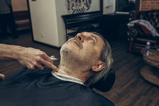 Close-up side profile portrait of handsome senior bearded caucasian man getting beard grooming in modern barbershop. Hairdresser serving client, making beard haircut using shaving machine. Barber shop concept.