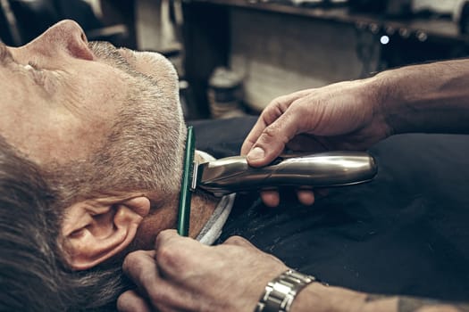 Close-up side profile portrait of handsome senior bearded caucasian man getting beard grooming in modern barbershop. Hairdresser serving client, making beard haircut using shaving machine. Barber shop concept.
