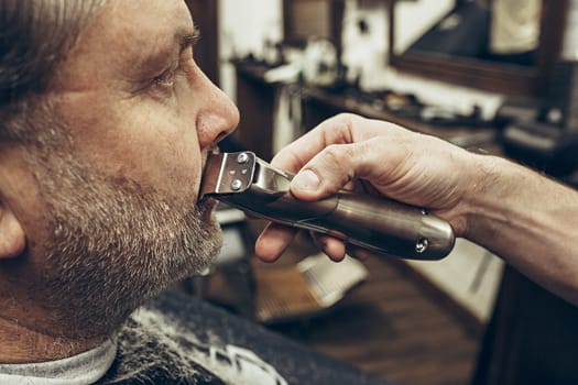 Close-up side profile portrait of handsome senior bearded caucasian man getting beard grooming in modern barbershop. Hairdresser serving client, making beard haircut using shaving machine. Barber shop concept.