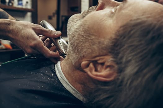 Close-up side back view handsome senior bearded caucasian man getting beard grooming in modern barbershop. Hairdresser serving client, making beard haircut using shaving machine. Barber shop concept.