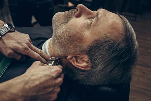 Close-up side back view handsome senior bearded caucasian man getting beard grooming in modern barbershop. Hairdresser serving client, making beard haircut using shaving machine. Barber shop concept.