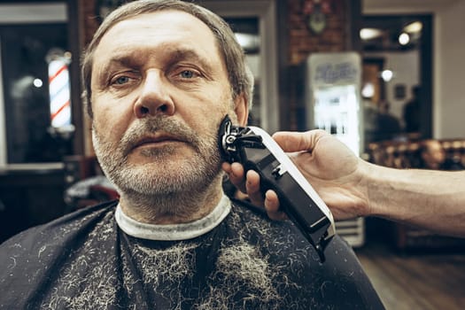 Close-up side view portrait of handsome senior bearded caucasian man getting beard grooming in modern barbershop. Hairdresser serving client, making beard haircut using shaving machine. Barber shop concept.