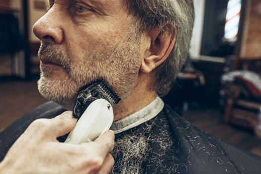 Close-up side view portrait of handsome senior bearded caucasian man getting beard grooming in modern barbershop. Hairdresser serving client, making beard haircut using shaving machine. Barber shop concept.