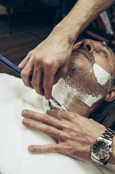 Close-up side top view handsome senior bearded caucasian man getting beard grooming in modern barbershop. Hairdresser serving client, making beard haircut using straight razor. Barber shop concept.