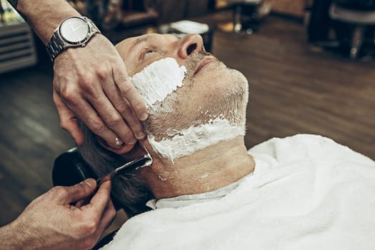 Close-up side top view handsome senior bearded caucasian man getting beard grooming in modern barbershop. Hairdresser serving client, making beard haircut using straight razor. Barber shop concept.