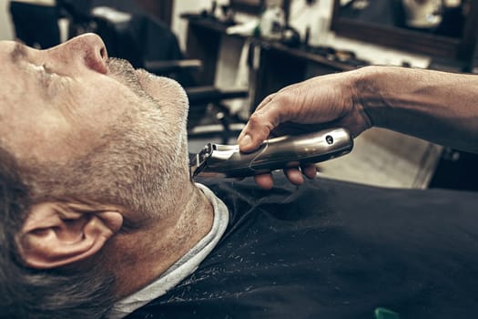 Close-up side profile portrait of handsome senior bearded caucasian man getting beard grooming in modern barbershop. Hairdresser serving client, making beard haircut using shaving machine. Barber shop concept.
