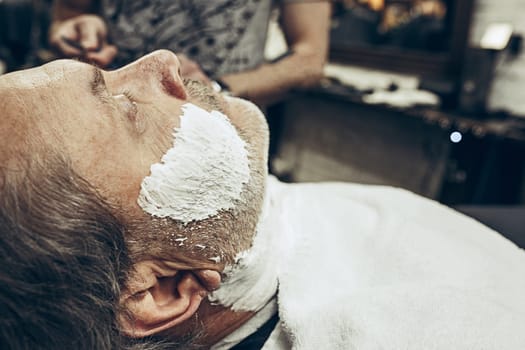 Close-up side view portrait of handsome senior bearded caucasian man getting beard grooming in modern barbershop. Hairdresser serving client, making beard haircut using straight razor. Barber shop concept.
