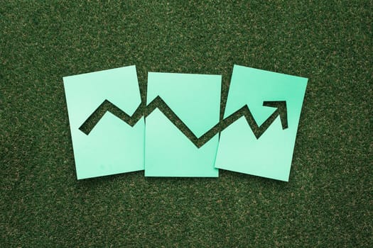 Successful financial graph with arrow pointing up composed of green paper cuts on grass, environment and business concept