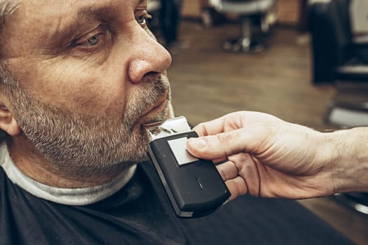 Close-up side view portrait of handsome senior bearded caucasian man getting beard grooming in modern barbershop. Hairdresser serving client, making beard haircut using shaving machine. Barber shop concept.