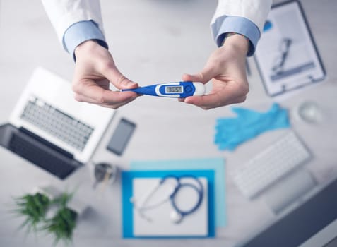 Doctor measuring temperature and holding a digital thermometer, desktop with medical equipment and computer on background, top view