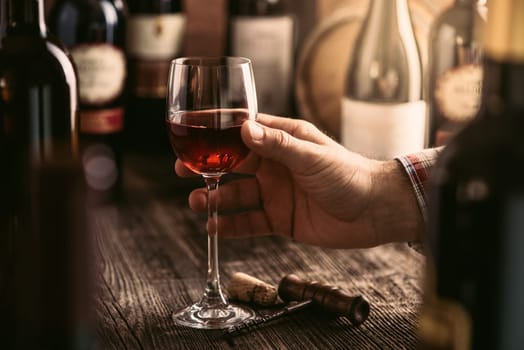 Wine tasting experience in the rustic cellar and wine bar: sommelier holding a glass of delicious red wine and excellent wine bottles collection on the background