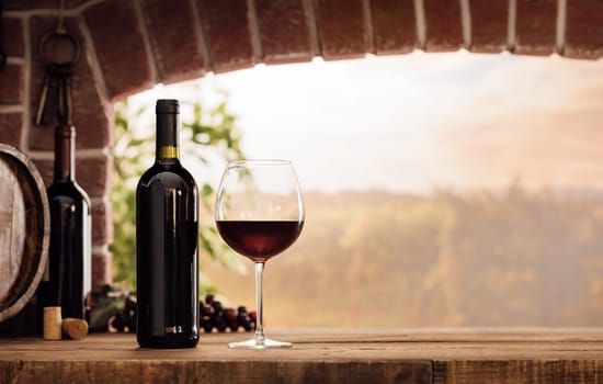 Red wine tasting in the wine cellar: wineglass and bottles next to the window and panoramic view of vineyards at sunset