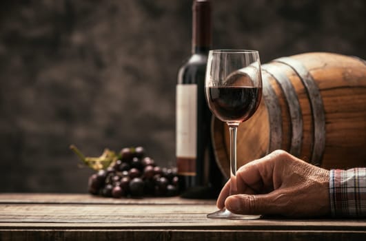 Senior wine maker tasting a glass of red wine in his cellar, wine bottle and wooden barrel on the background, wine tradition and culture concept