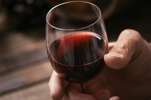Senior man tasting a glass of italian red wine, rustic wooden table on the background, hand close up