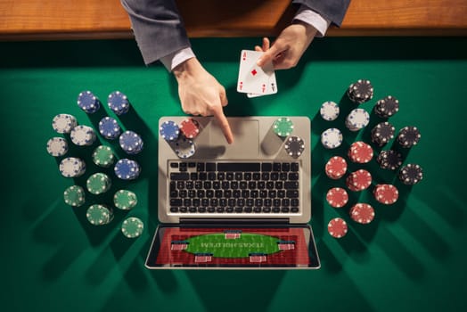 Online player's hands with laptop and stack of chips all around on green table top view, he is holding two ace cards