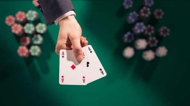 Elegant male poker player holding two aces with stacks of chips on background and green table, top view