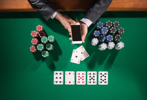 Elegant male poker player using a smartphone, cards and chips stacks all around, green table top view
