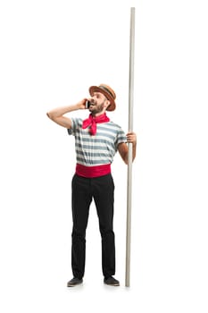 Caucasian funny bearded man in traditional gondolier costume and hat with paddle standing with mobile phone at studio isolated on white background. young man. Human emotions, facial expression concept