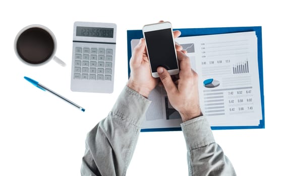 Businessman at desk using a touch screen smart phone, calculator and financial report, hands top view