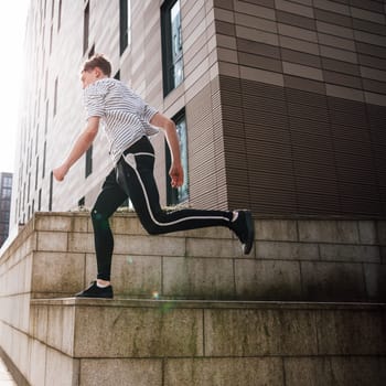 Young freerunner is doing stunts in the city. He is running along a building wall in the sun.