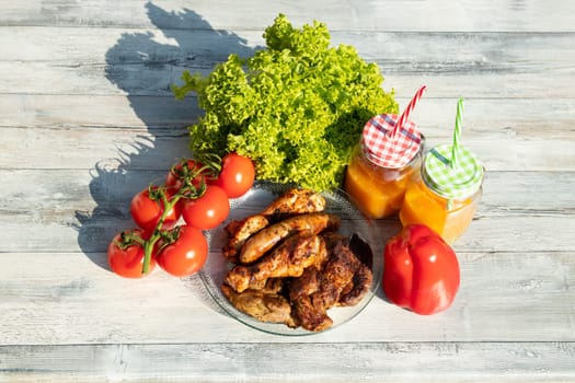 Top view on plate of fresh grilled meat surrounded by fresh salad, tomatoes, paprika and two glasses of freshly squeezed orange juice. presented outdoor on wooden table.