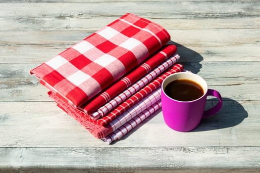 Stack of red white checkered and striped linen tableclothes and a cup of coffee on rustic bright wooden table. Outdoor.
