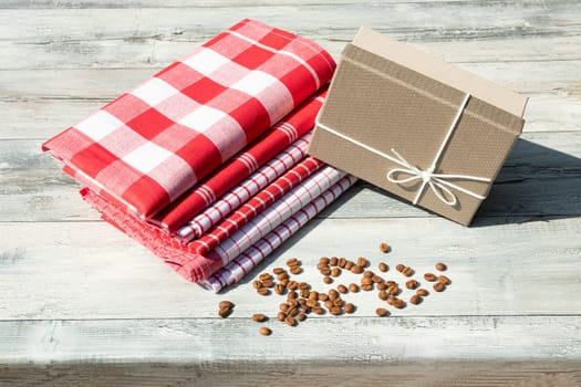 Greeting card. Birthday gift box with stack of tableclothes and roasted coffee beans on rustic bright wooden table. Outdoor. Card concept.
