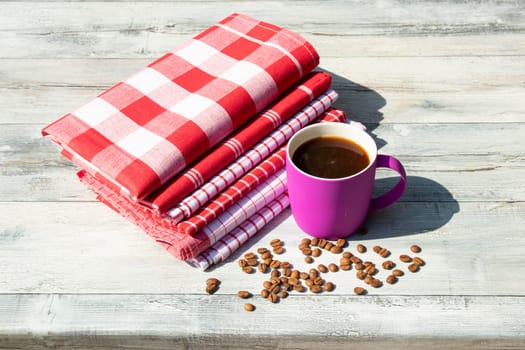 Purpure cup of hot black coffee with roasted coffee beans together with a Stack of red white checkered and striped linen tableclothes on rustic bright wooden table. Outdoor