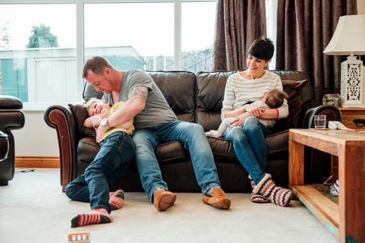 Couple are relaxing at home with their children. The mother is breastfeeding the baby while watching her husband and son playfight.