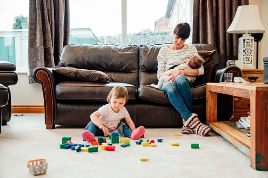 A mother is watching her daughter play with toy blocks in the living room as she breastfeeds the baby on the sofa.