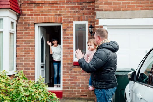 Father is carrying his baby daughter to the car and is turning round to wave at the mother who is stood at the front door of the house.
