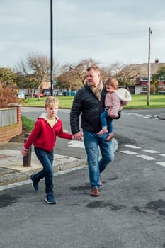 Mature man is crossing the road with his children, his son is holding his hand and he is carrying his daughter on his hip.